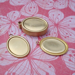 May include: Three gold-colored oval-shaped metal pendant trays with a smooth surface. The trays are different sizes and have a loop at the top for hanging.