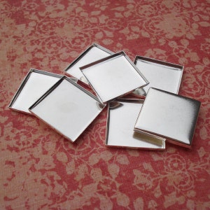 May include: A group of six silver square metal trays, each with a smooth, reflective surface. The trays are arranged in a random pattern on a red floral fabric background.