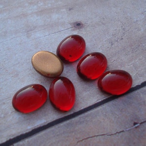 May include: Six red oval glass cabochons and one gold oval cabochon. The red cabochons are smooth and shiny. The gold cabochon is dull and has a slightly textured surface.