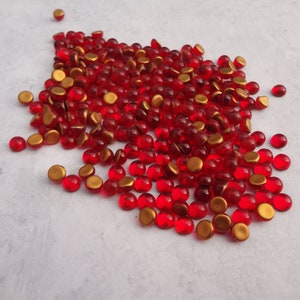 May include: A large pile of red glass cabochons with gold foil backs. The cabochons are round and have a smooth, shiny surface.