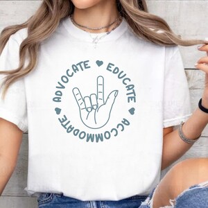 Puede incluir: Camiseta blanca con un gráfico turquesa. El gráfico presenta una mano en el signo de 'Te amo', rodeada por las palabras 'Advocate, Educate, Accommodate' y pequeños símbolos de corazón. La camiseta es de material suave.