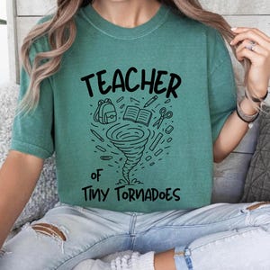 Op de afbeelding: Teal groen t-shirt met de woorden "TEACHER of TINY TORNADOES" in zwart schrift. Een zwart-witte afbeelding van een tornado met schoolspullen, waaronder een rugzak, een boek, een schaar en potloden, staat in het midden van het shirt.