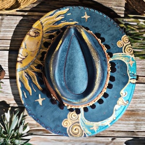 May include: A teal cowboy hat with a celestial design. The hat features a dark teal crown and a wide brim decorated with a yellow sun, a crescent moon, stars, and gold accents. The brim is trimmed with small, light-colored beads.