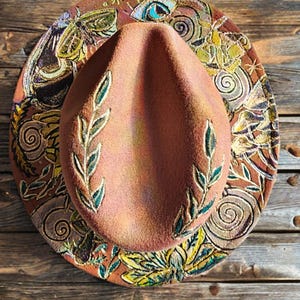 May include: A brown fedora-style hat with a decorative brim. The hat features embroidered green leaves on the crown and a patterned brim with floral and insect designs in shades of brown, yellow, and green. The hat is displayed on a wooden surface.