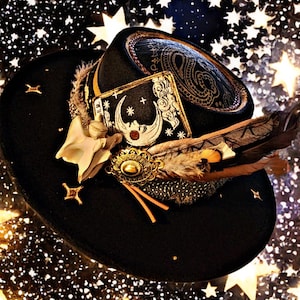 May include: A black hat with a decorative brim, adorned with a gold-colored embellishment, feathers, and a card featuring a crescent moon design. The hat also has a star-shaped accent and small gold studs.