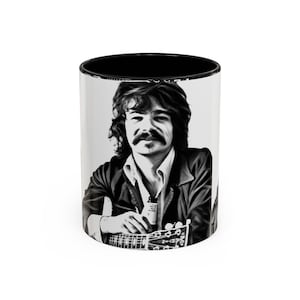 May include: A white ceramic coffee mug with a black interior. The mug features a black and white portrait of a man with a mustache holding a guitar. The mug is a standard 11 oz size.