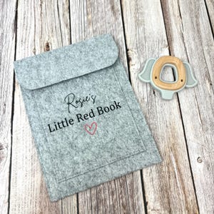 May include: A gray felt book cover with the text "Rosie's Little Red Book" and a small red heart. A light blue wooden teether shaped like an elephant is in the background.