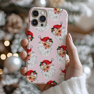 May include: A pink phone case featuring a repeating pattern of vintage Santa Claus faces. Each Santa has a red hat, white beard, and rosy cheeks. The case is held in front of a blurred Christmas tree.