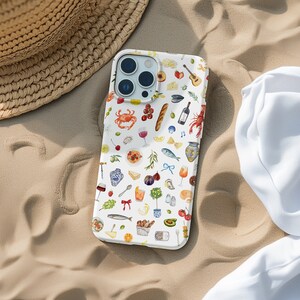 Puede incluir: Funda blanca para iPhone con un patr&oacute;n colorido de ilustraciones de comida y bebida, incluyendo un cangrejo, vino, pan y una botella de vino. La funda est&aacute; sobre arena, junto a un sombrero de paja y una tela blanca.