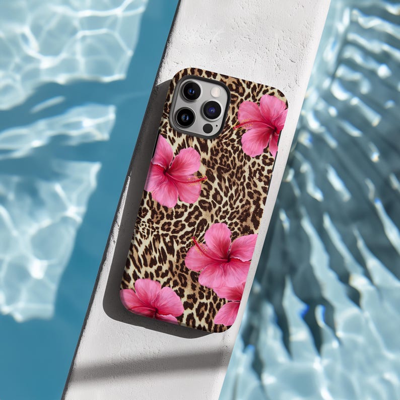 May include: A phone case featuring a leopard print background with pink hibiscus flowers. The case has a camera lens cutout and a black border.