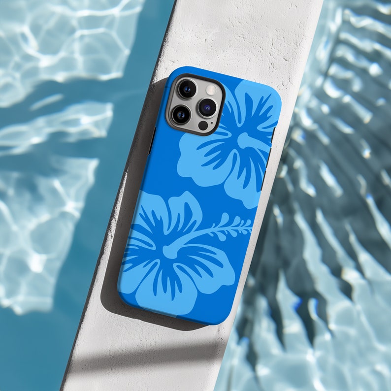 Op de afbeelding: Een blauwe iPhone-hoes met een bloemenpatroon met twee witte hibiscusbloemen. De hoes heeft een uitsparing voor de cameralens en een witte achtergrond.