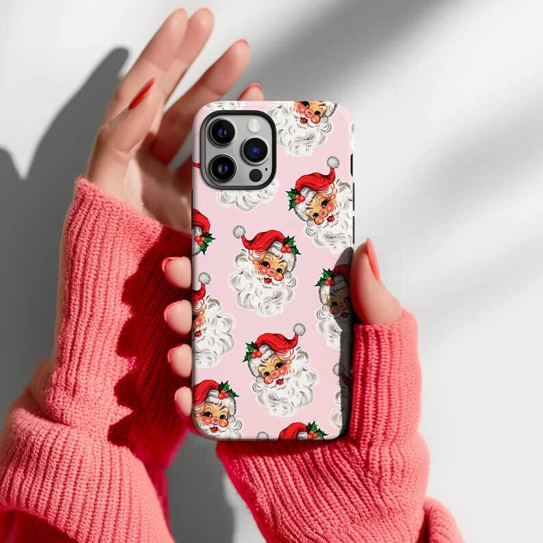 May include: A pink phone case featuring a repeating pattern of vintage Santa Claus faces with red hats, white beards, and holly accents. The case is held by a person wearing a pink sweater.