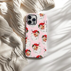 May include: A pink phone case featuring a repeating pattern of vintage Santa Claus faces. Each Santa has a red hat, white beard, and a sprig of holly. The case is shown on a white knitted surface.