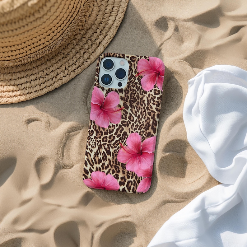 May include: A phone case with a leopard print background and pink hibiscus flowers. The case is lying on a sandy surface.