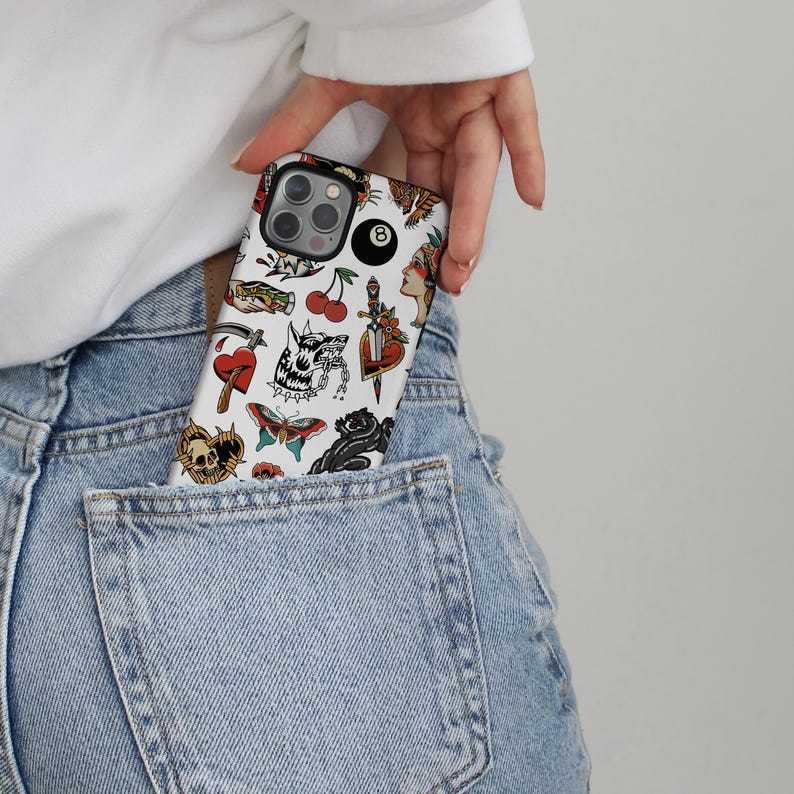 May include: A white phone case with a variety of colorful tattoo-style designs, including a skull, a heart, a butterfly, a cherry, a dagger, and a tiger. The case also features a black 8-ball.