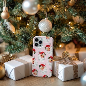May include: A pink phone case featuring a repeating pattern of Santa Claus faces with red hats and white beards. The case is displayed in front of a Christmas tree with ornaments and wrapped gifts.