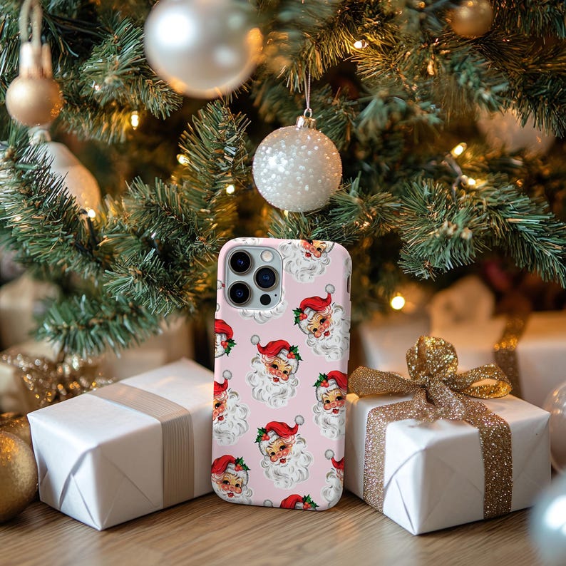May include: A pink phone case featuring a repeating pattern of Santa Claus faces with red hats and white beards. The case is displayed in front of a Christmas tree with ornaments and wrapped gifts.