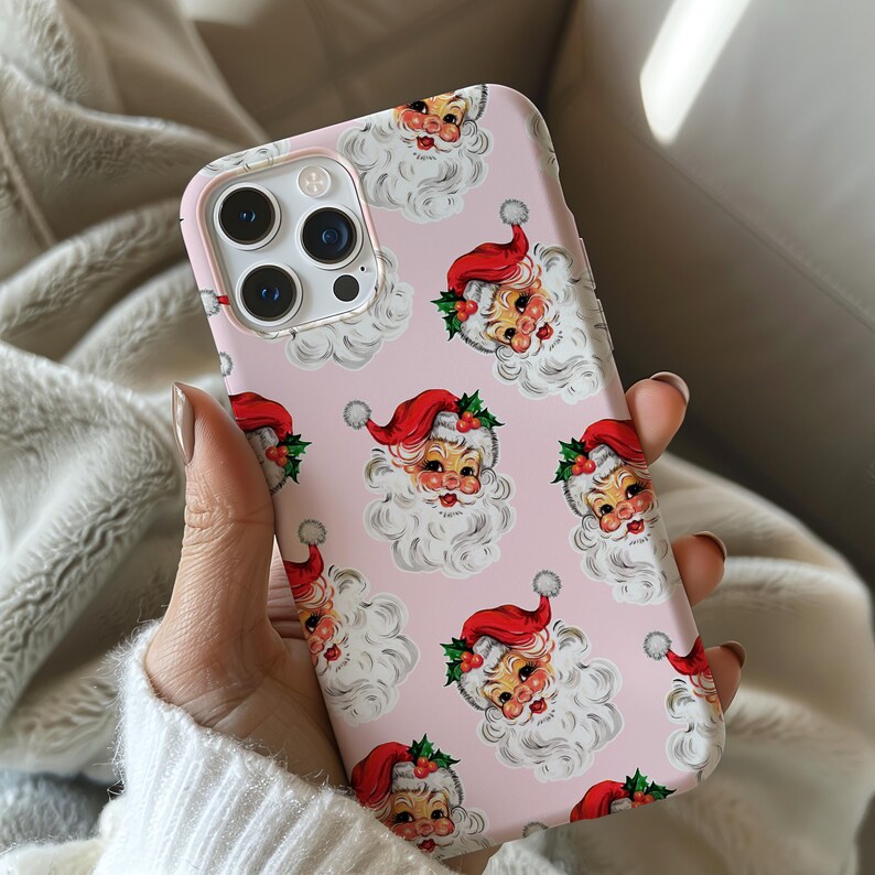 May include: A pink phone case featuring a repeating pattern of Santa Claus faces with red hats and white beards. The case has a light pink border and a camera cutout.
