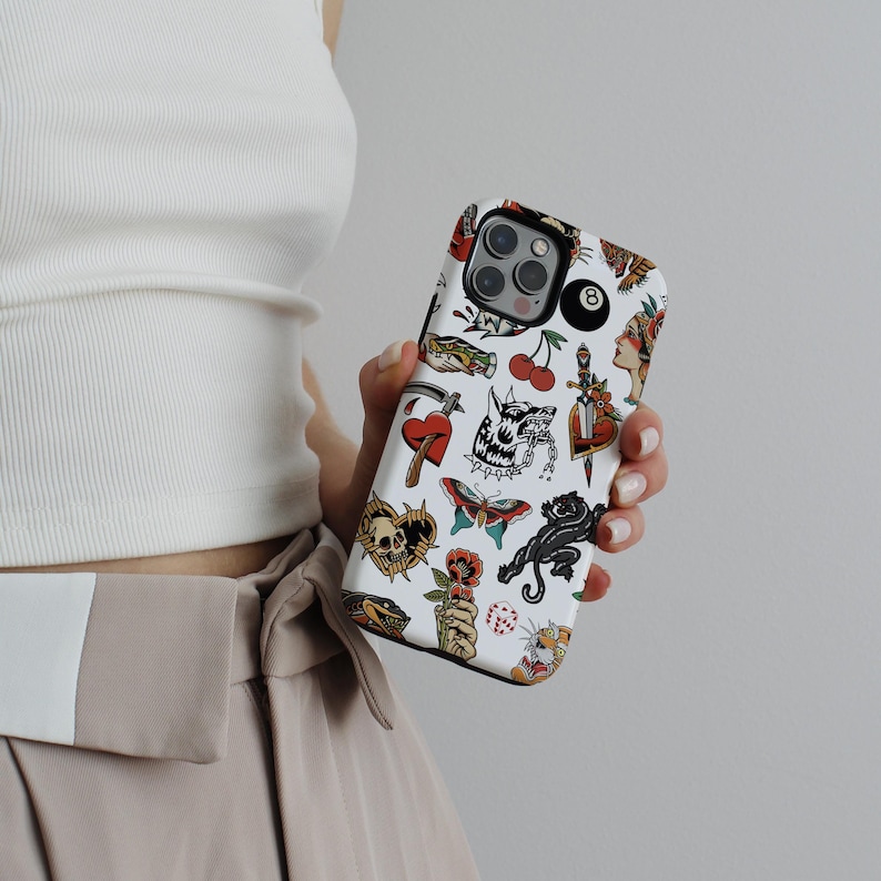May include: White iPhone case with a colorful tattoo-style design featuring a skull, a panther, a heart, a dagger, a cherry, a dice, and a 8-ball. The design is on a white background with black outlines.