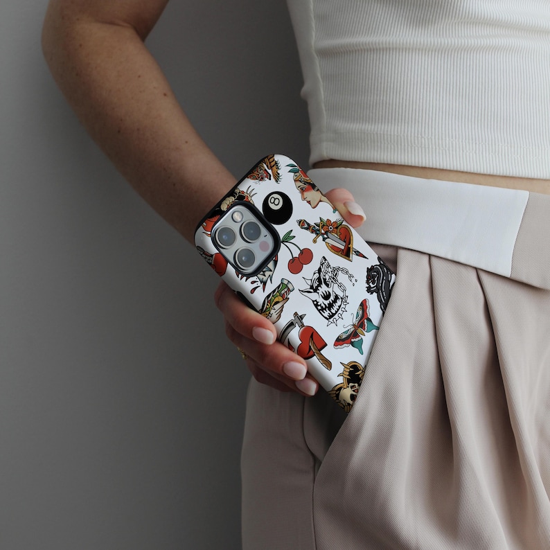 May include: A white iPhone case with a colorful tattoo-style design. The design includes a skull, a heart, a dagger, a butterfly, cherries, and a pool ball. The case is being held by a person's hand.