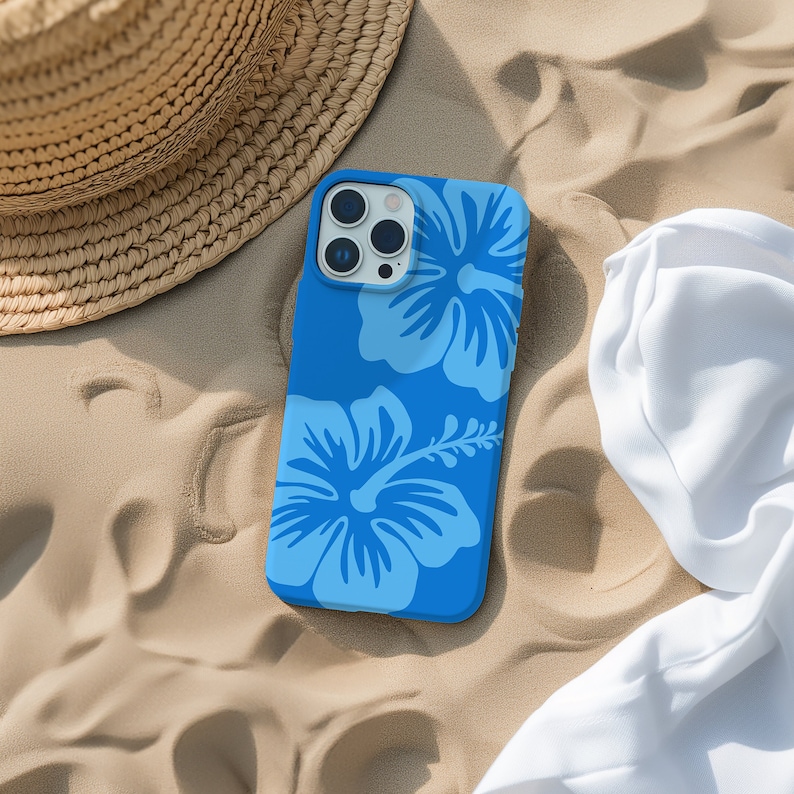 Op de afbeelding: Een blauwe telefoonhoes met een bloemenpatroon met hibiscusbloemen. De hoes ligt op een zanderig oppervlak.