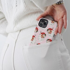 May include: A pink phone case featuring a repeating pattern of Santa Claus faces with red hats and white beards. The phone case is in a white pocket, with a white sweater and white pants in the background.