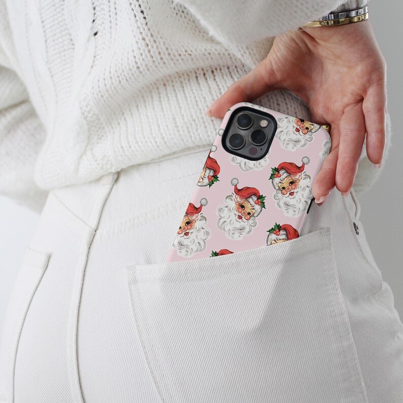 May include: A pink phone case featuring a repeating pattern of Santa Claus faces with red hats and white beards. The phone case is in a white pocket, with a white sweater and white pants in the background.