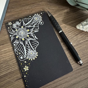 May include: A black notebook with a floral design in white and yellow ink. A black pen rests on the notebook, which is on a wooden surface. The floral design is on the top left corner of the notebook.