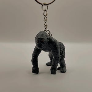 May include: A black gorilla keychain with a silver chain and keyring. The gorilla is standing on all fours, with detailed fur texture and red eyes. The keychain is a novelty item.