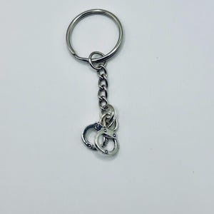 May include: Silver-toned keychain featuring a small pair of handcuffs. The handcuffs are attached to a chain and a large key ring. The keychain is made of metal and has a shiny finish.