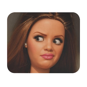 Peut inclure: Une tête de poupée aux longs cheveux bruns, sourcils arqués et longs cils. La poupée a une expression faciale sceptique avec les yeux tournés sur le côté et des lèvres roses. Une petite boucle d'oreille à clou argentée est visible.