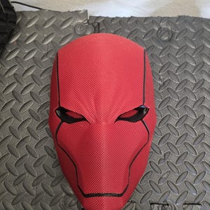 Puede incluir: Una máscara roja texturizada con aberturas negras para los ojos y contornos negros. La máscara tiene un diseño distintivo, con una barbilla puntiaguda y una línea de cejas definida. La máscara es probablemente para cosplay o disfraces.