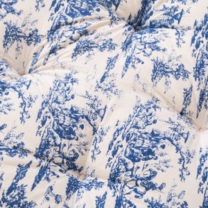 May include: A white fabric cushion with a blue and white toile pattern. The fabric has a quilted texture and is decorated with small buttons.