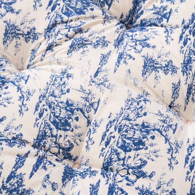 May include: A white fabric cushion with a blue and white toile pattern. The fabric has a quilted texture and is decorated with small buttons.