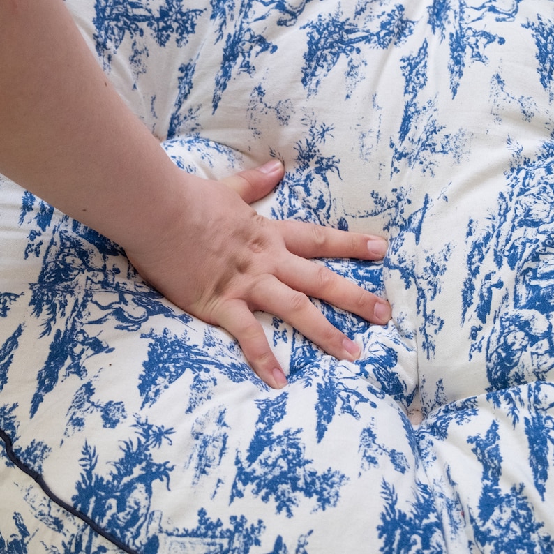May include: A white and blue patterned cushion with a hand resting on top. The cushion has a blue and white floral design.