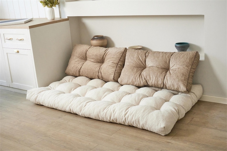 May include: A floor cushion set with two square back pillows and a rectangular base cushion. The cushions are beige and cream-colored, with a tufted design. The set is placed against a white wall and a light-colored wooden floor.