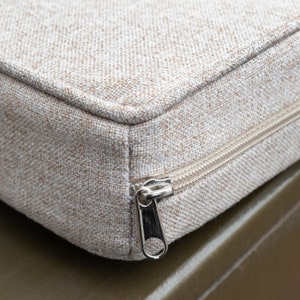 May include: Close-up of a beige zippered pouch with a silver zipper pull. The pouch is made of a textured fabric.