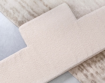 May include: A textured, off-white, T-shaped rug piece with a dense, looped pile surface. This solid-colored textile segment is layered over a larger, rectangular rug featuring a subtle, mottled beige and cream abstract pattern on a flat surface.