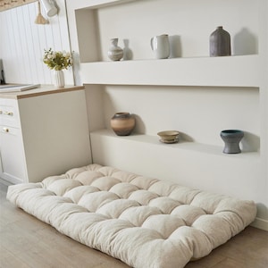 May include: A rectangular floor cushion featuring a tufted, off-white textured fabric surface. The cushion is thick and plush, with a grid of circular indentations across its top, providing a soft, padded base for sitting or lounging on the floor.