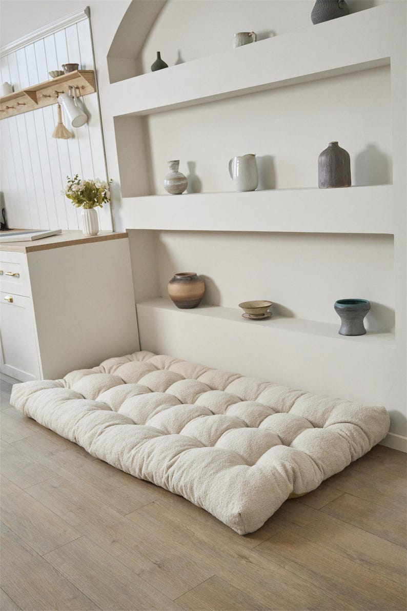 May include: A rectangular floor cushion featuring a tufted, off-white textured fabric surface. The cushion is thick and plush, with a grid of circular indentations across its top, providing a soft, padded base for sitting or lounging on the floor.