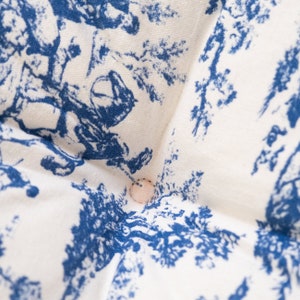 May include: A white fabric cushion with a blue and white toile pattern. The cushion has a button in the center.