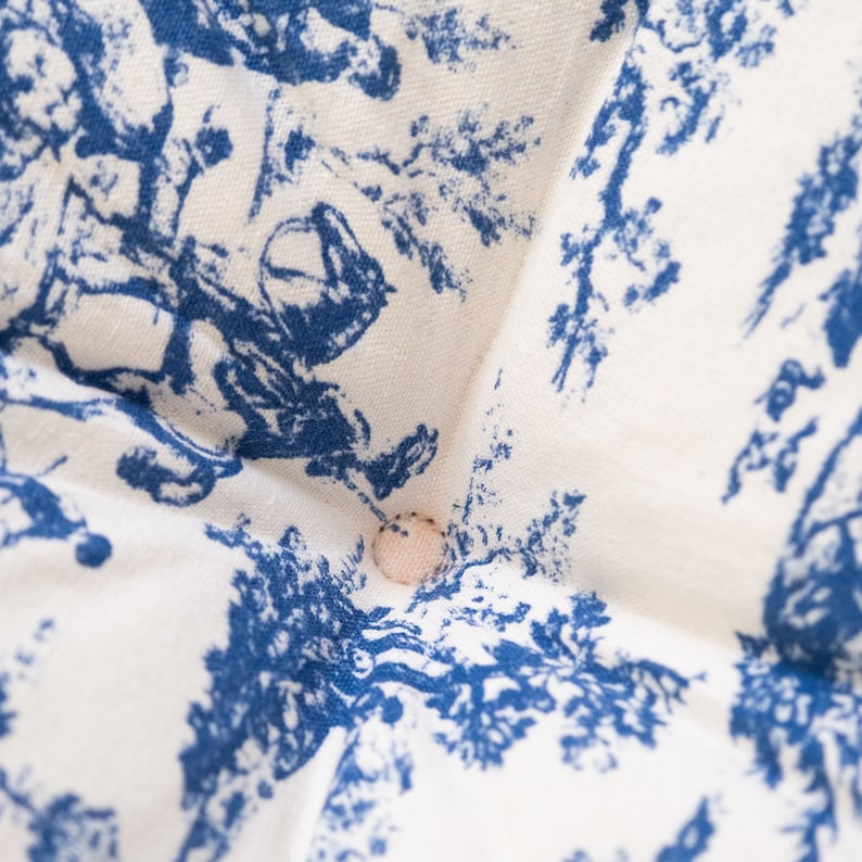 May include: A white fabric cushion with a blue and white toile pattern. The cushion has a button in the center.