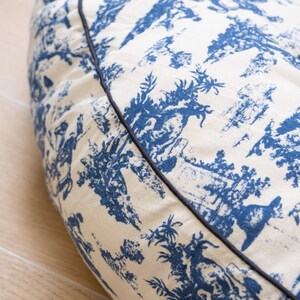 May include: A round, cream-colored dog bed with a blue and white toile pattern. The bed has a black trim around the edge.