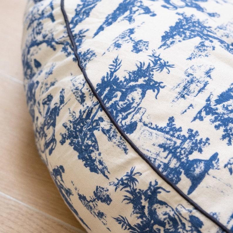 May include: A round, cream-colored dog bed with a blue and white toile pattern. The bed has a black trim around the edge.