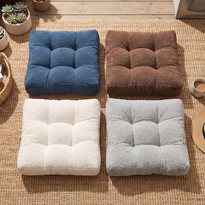 May include: A set of four square floor cushions featuring a plush, textured sherpa-style fabric. The cushions are arranged in a grid pattern and come in four distinct solid colors: deep blue, warm brown, off-white, and light gray.