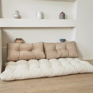 May include: A floor cushion featuring a tufted off-white base and two rectangular tan back cushions. The base is thick and rectangular with a grid of tufted indentations, while the two back cushions are placed upright against the wall.