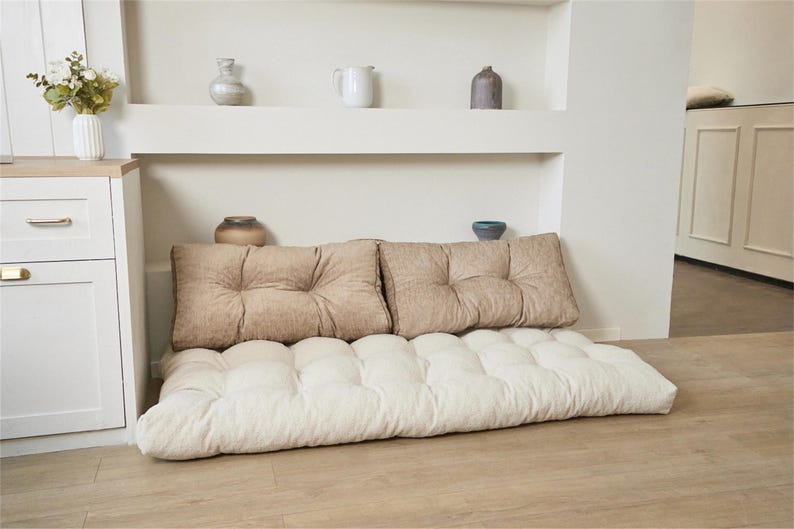 May include: A floor cushion featuring a tufted off-white base and two rectangular tan back cushions. The base is thick and rectangular with a grid of tufted indentations, while the two back cushions are placed upright against the wall.