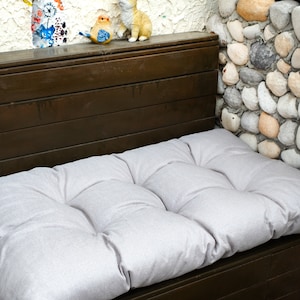 May include: A rectangular, light gray tufted bench cushion with a textured fabric surface. The cushion is positioned on a dark brown wooden bench, featuring a series of horizontal slats and a flat top surface for seating comfort.