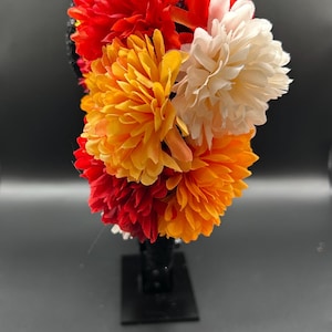 May include: A headband featuring a cluster of artificial flowers in vibrant shades of red, orange, and cream. The base of the headband is black and decorated with small, reflective sequins that catch the light, providing a contrast to the petals.