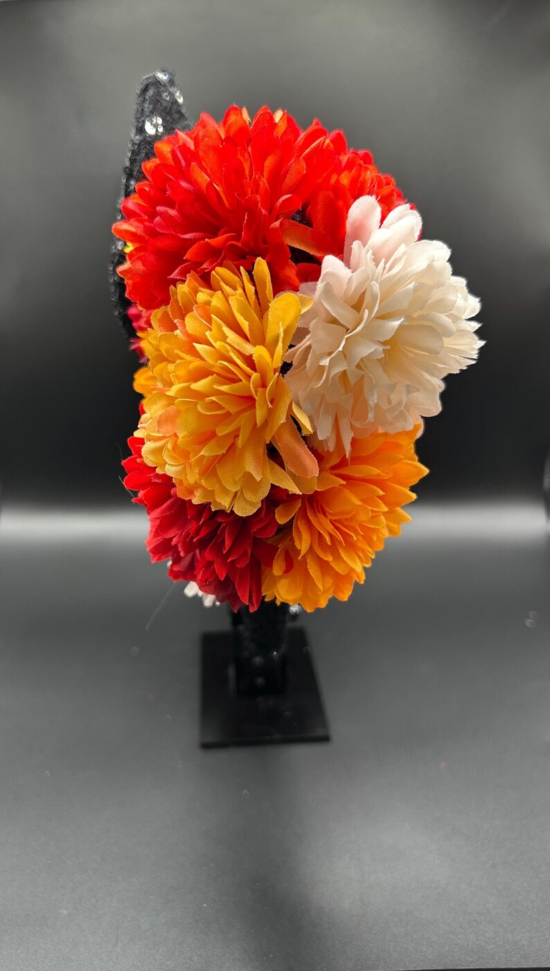 May include: A headband featuring a cluster of artificial flowers in vibrant shades of red, orange, and cream. The base of the headband is black and decorated with small, reflective sequins that catch the light, providing a contrast to the petals.